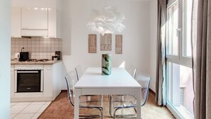 A modern kitchen with white cabinets, a built-in oven, and a dining area with a white table and clear chairs. - App. near italian border (Pink Flat 2) (Chiasso)
