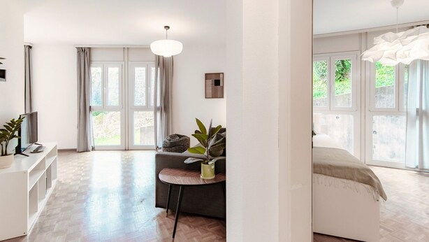 A modern hotel room with a large window, a sofa, a small round table, and a bed. - App. near italian border (Pink Flat 2) (Chiasso)