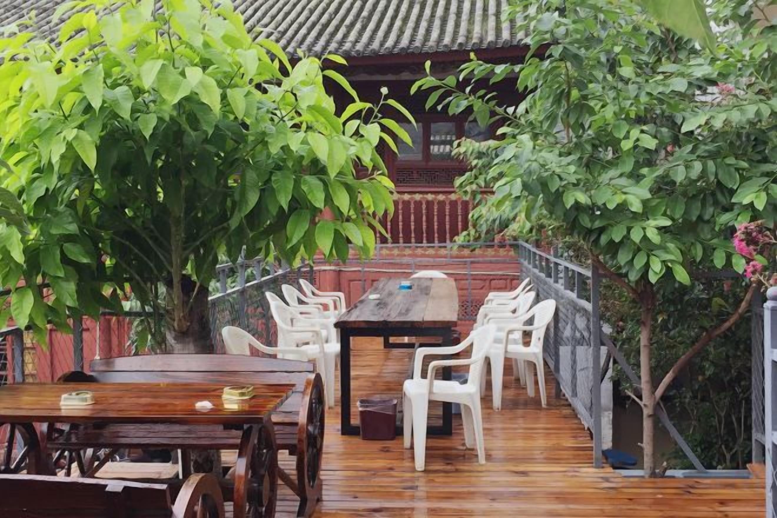 An outdoor wooden deck with a dining table and chairs, surrounded by greenery and a traditional building in the background.