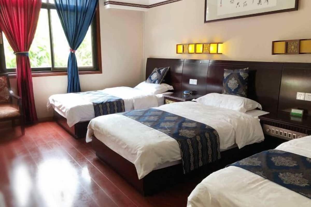 A hotel room with two single beds, a wooden floor, a window with curtains, and a wall-mounted light fixture.