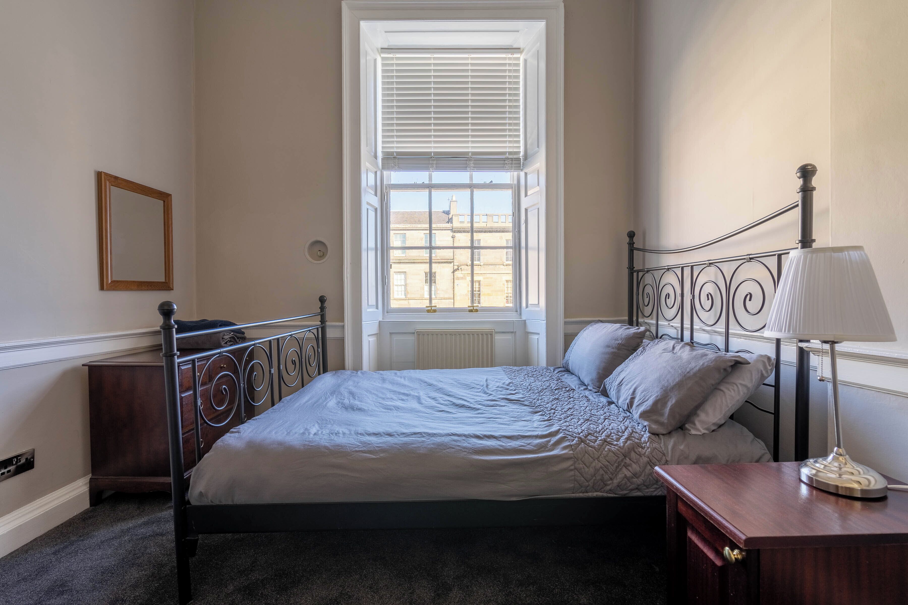 Una camera da letto con un letto, un comodino, una lampada, una finestra con persiane e un termosifone.