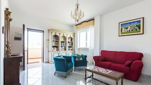 A living room with a red sofa, two blue armchairs, a wooden coffee table, and a chandelier. There is a bookshelf filled with books and a painting on the wall. - 4018 Margaret House by Barbarhouse (Fasano)