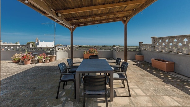 A covered outdoor seating area with a table and chairs, potted plants, and a view of the sea. - 4018 Margaret House by Barbarhouse (Fasano)