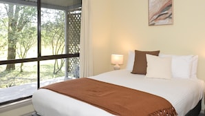 Un dormitorio con un ventanal grande, una cama con ropa de cama blanca y marrón, una lámpara y un cuadro en la pared.