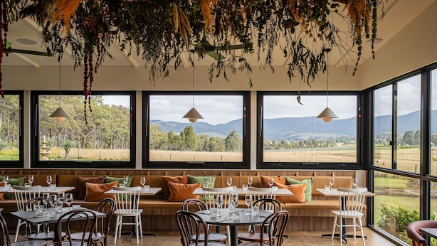 Un restaurante con amplios ventanales que ofrecen vistas al campo. Mesas y sillas dispuestas para comer.