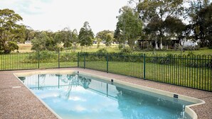 A rectangular swimming pool with a clear blue water surface, surrounded by a black metal fence and a pebble-textured deck. - Twin Tree Bush Cabin in the Heart of the Hunter Valley! 
1 of 10 Cabins listed! (Pokolbin)