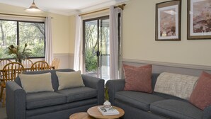 A living room with a grey sofa, two wooden side tables, and a dining area with wooden chairs and a table. - Twin Tree Bush Cabin in the Heart of the Hunter Valley! 
1 of 10 Cabins listed! (Pokolbin)