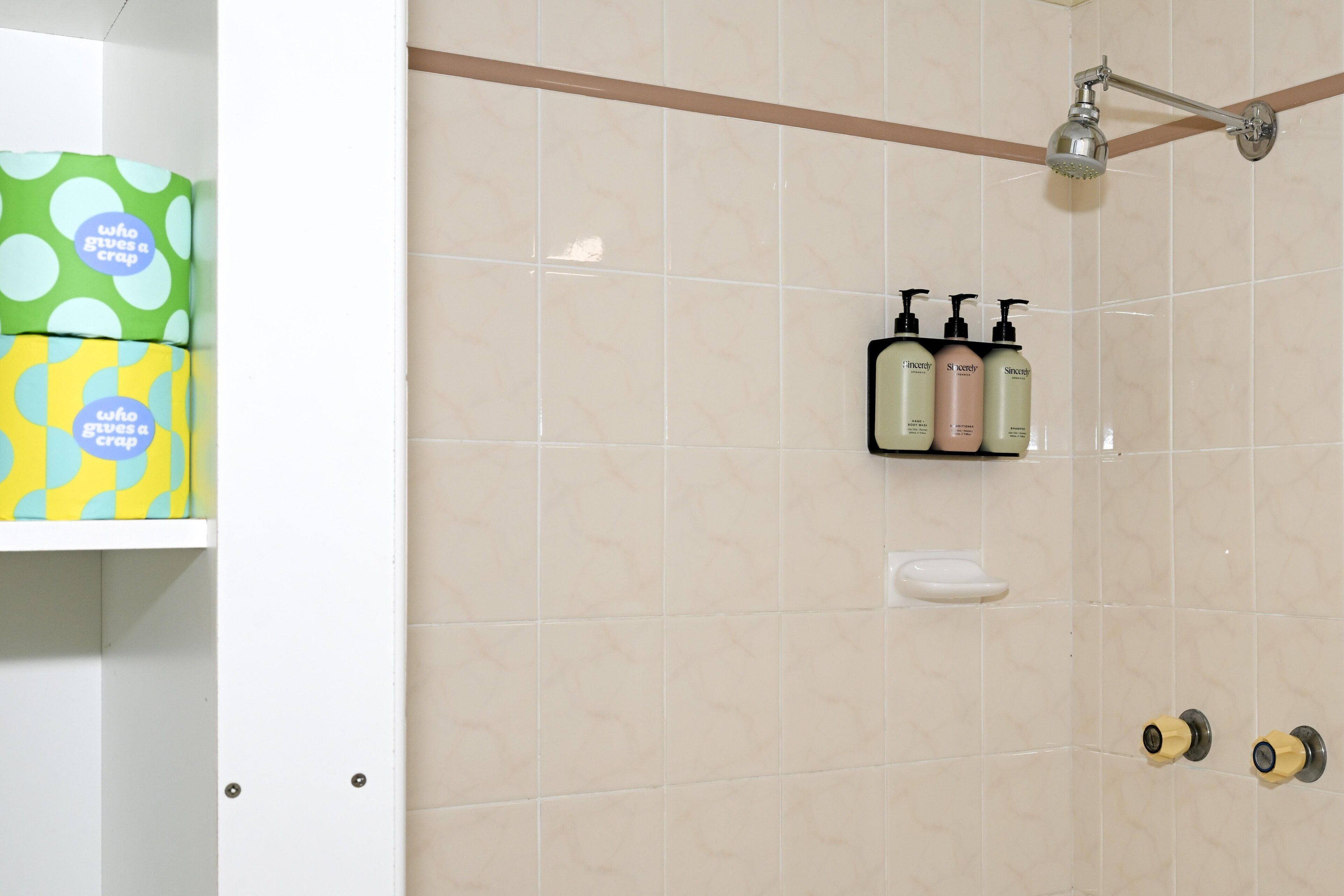 A bathroom with beige tiles, a showerhead, and toiletries on a shelf.