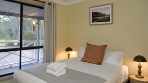 Una cama bien hecha con una colcha gris, almohadas blancas y un cojín decorativo marrón. Hay dos lámparas de noche sobre mesitas de madera. Un cuadro enmarcado cuelga en la pared sobre la cama.