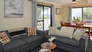 A modern living room with a grey sofa set, a round wooden coffee table, and a large window with curtains. - Cedar Cabin 1 - 2 bedroom with 1 x queen and 1 King or 2 king singles + sofa bed (Pokolbin)