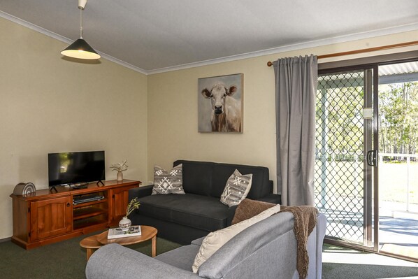 A living room with a black sofa, a wooden entertainment unit, a television, and a painting of a cow on the wall. - Callistemon Cabin 2 - 2 bedroom with 1 x queen and 1 King or 2 king singles + sofa bed (Pokolbin)
