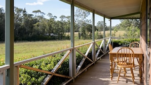 A covered outdoor area with a wooden table and chairs, surrounded by a metal railing, overlooking a grassy field and trees.