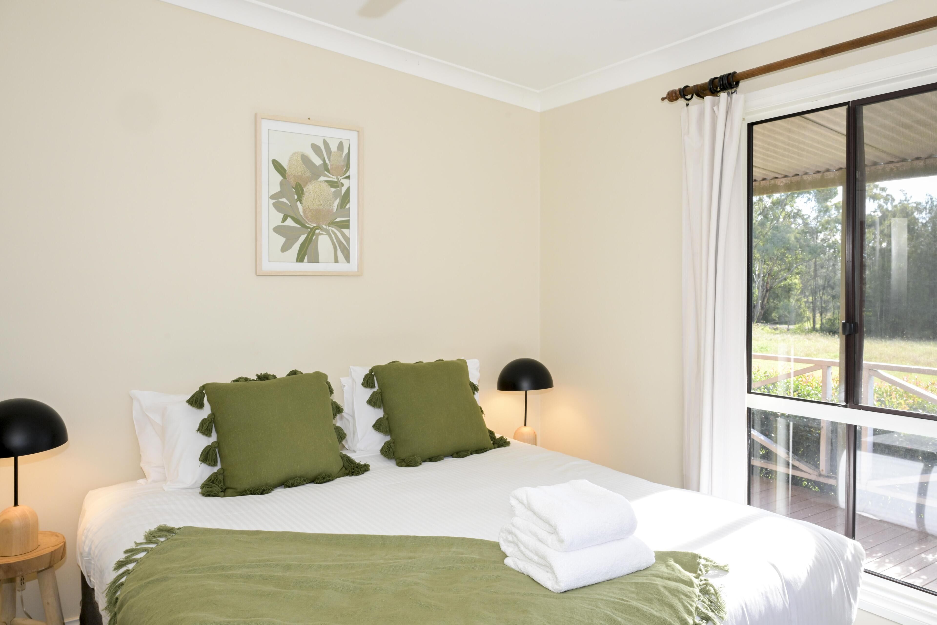 Un dormitorio con una cama, dos almohadas, una lámpara de noche, un cuadro enmarcado y una ventana con vista a follaje.