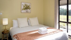 Una cama bien tendida con sábanas blancas y un edredón color melocotón, una lámpara de noche e impresión enmarcadas de motivos botánicos en la pared.