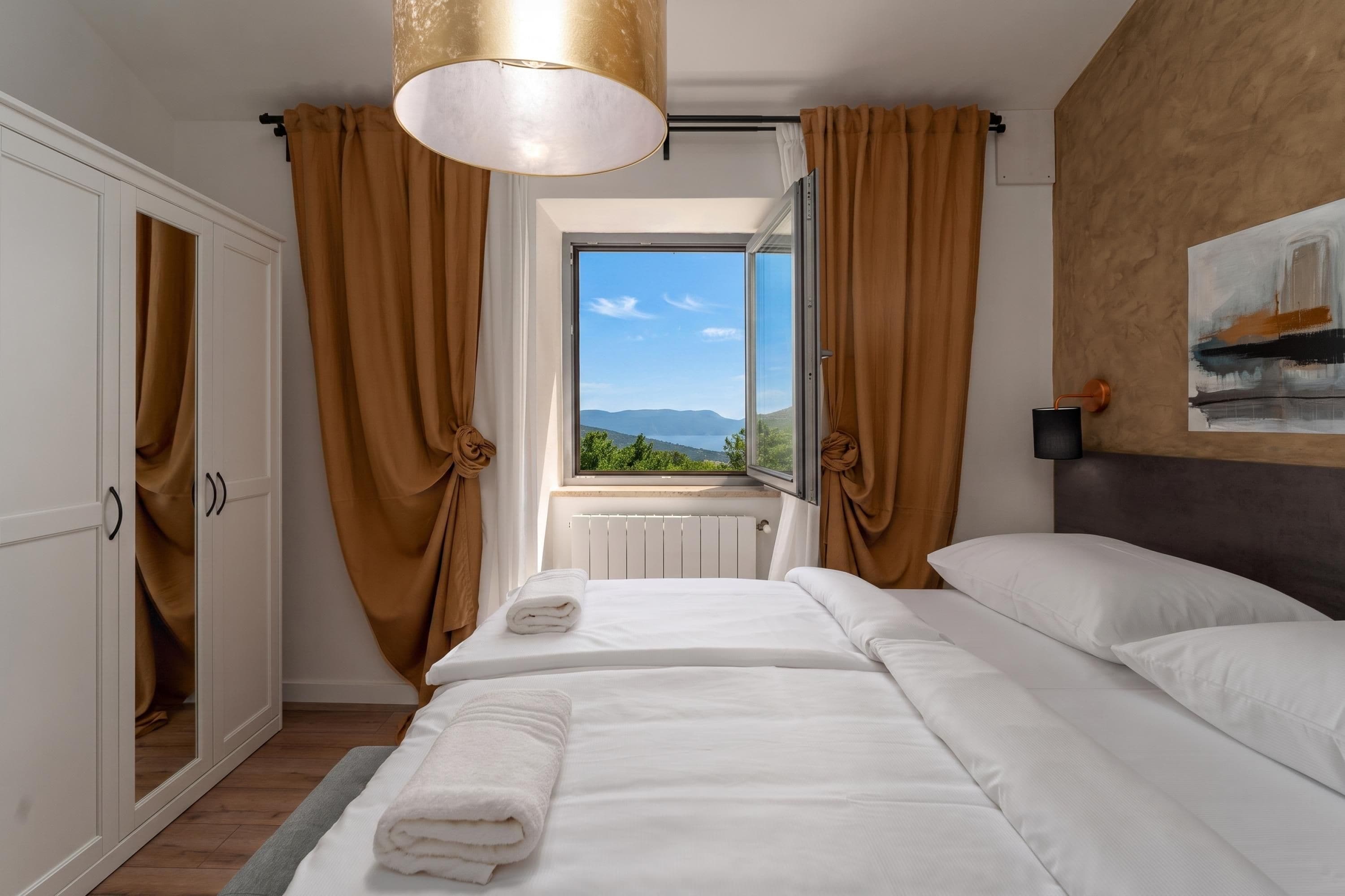 A hotel room with a bed, a window with a view, a radiator, and a painting on the wall.