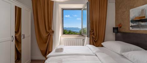 A hotel room with a bed, a window with a view, a radiator, and a painting on the wall.