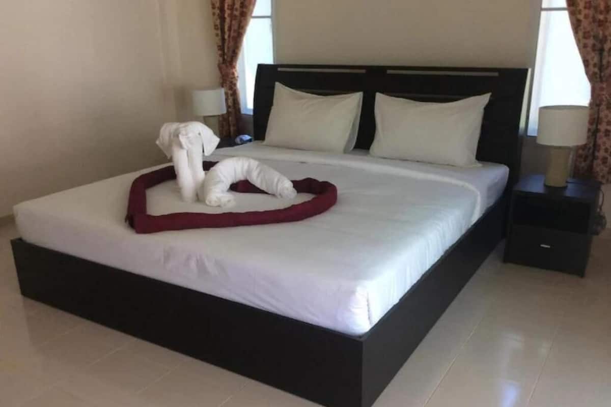 Cama bem arrumada com lençóis brancos e um travesseiro decorativo vermelho. Tem uma mesinha de cabeceira pequena com uma luminária e uma gaveta.