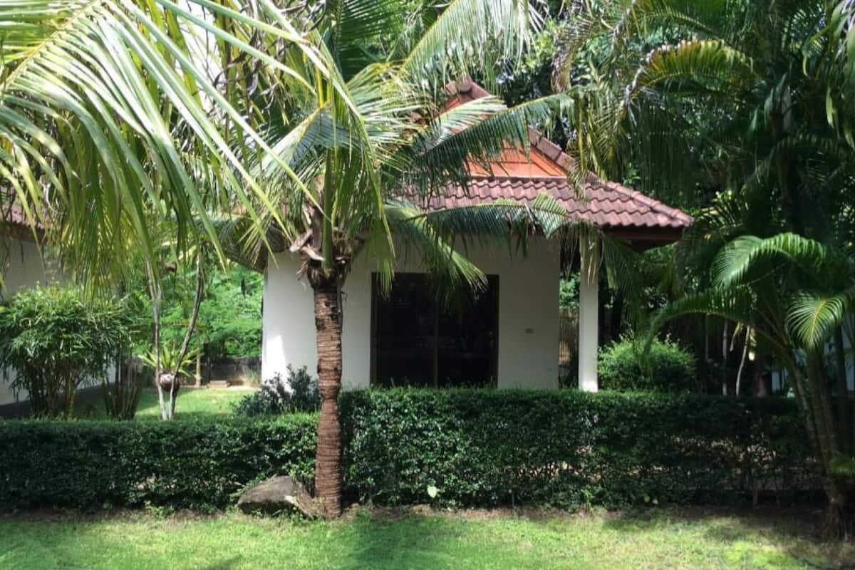Um pequeno edifício com telhado de cerâmica, cercado por vegetação exuberante e palmeiras.