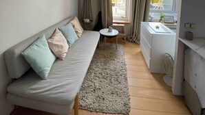 A compact living space with a sofa, a small table, a window with curtains, and a kitchenette area.