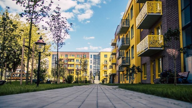 A pathway leading to a row of multi-story buildings with balconies, trees, and benches.