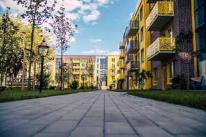 Un chemin qui mène à une rangée d’immeubles à plusieurs étages, dotés de balcons, d’arbres et de bancs.