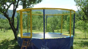 A circular trampoline with a yellow and blue safety net, surrounded by grass and trees. - Roulotte Cévenole (Aulas)
