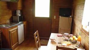 A small kitchen with wooden cabinets, a sink, a microwave, and a window. There is a table with chairs, a bowl of fruit, and a bottle of juice. - Roulotte Cévenole (Aulas)