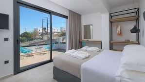 A modern hotel room with a large sliding glass door to a balcony, a bed with neatly folded towels, a wooden desk, and a hanging light fixture.