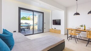 A modern living room with a sofa, a dining area with a table and chairs, a TV mounted on the wall, and a sliding glass door leading to an outdoor patio.