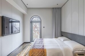 A modern hotel room with a large bed, a flat-screen TV mounted on the wall, and a view through a large arched window. - Xiongan Kapok Hotel (Xiong County)