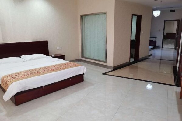 A hotel room with a large bed, a nightstand, a window with curtains, and a hallway leading to another room.
