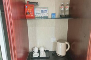 A hotel room with a shelf containing a box, a small clock, and bottled water. There is a coffee maker and a tray with cups and a teapot on the counter.