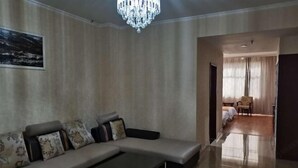 A modern living room with a large sectional sofa, a coffee table, and a chandelier. There is a painting on the wall and a view into another room with a bed and curtains. - Changshou Business Hotel (Luntai)