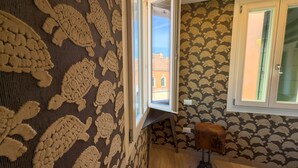 A room with patterned wallpaper, a window with a view of buildings, and a small stool. - Ca de la Corona by Ca-sa Check (Chioggia)