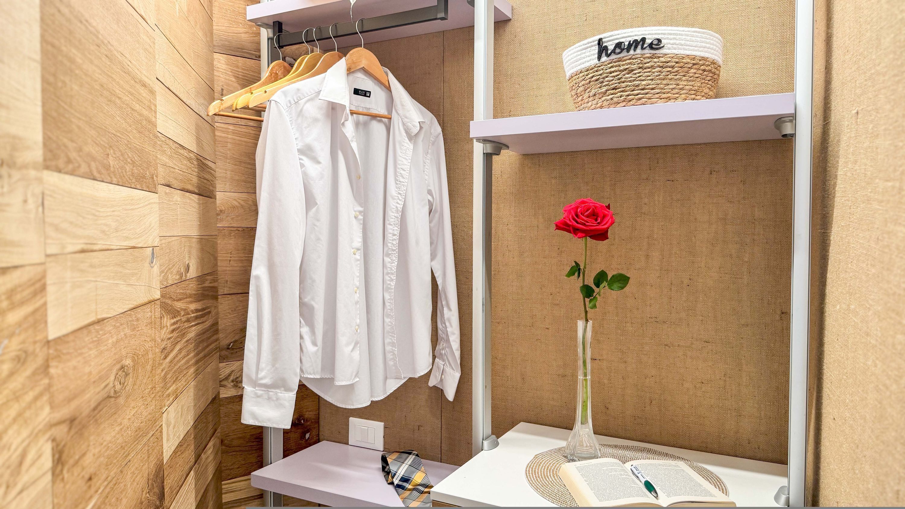 A neatly organized room with a wooden wall, a white shirt hanging on a wooden hanger, a clear vase with a single red rose, and a basket labeled "home" on a shelf.