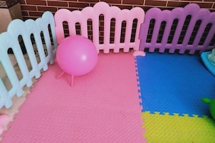 Zona de juegos con un balón rosa, una valla blanca y morada, y alfombras de espuma multicolores.