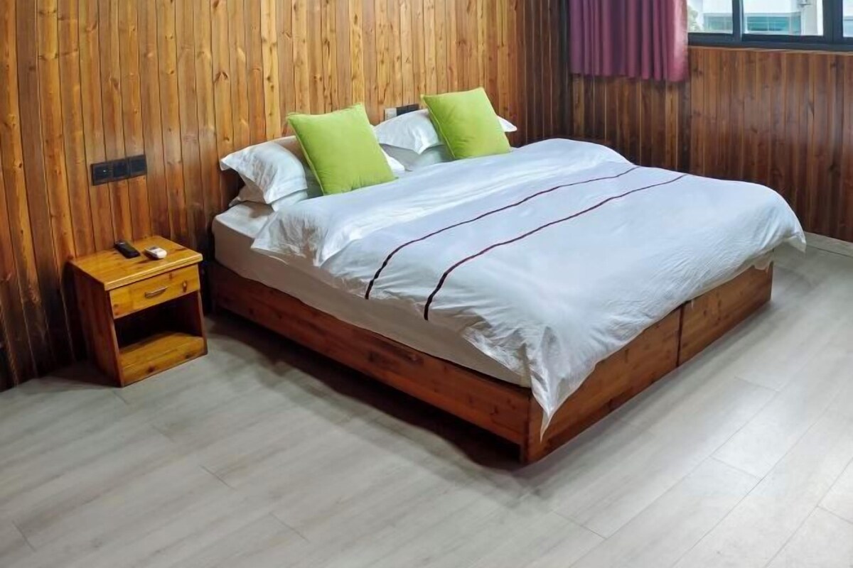 A wooden bed with a white and green bedspread, a nightstand with a phone and a small drawer, and a wooden wall with a window and curtains.