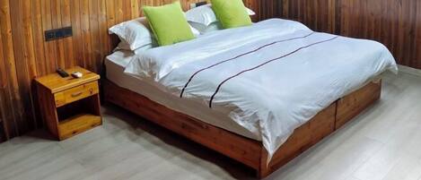 Una cama de madera con cubrecama blanco y verde, una mesita de noche con un teléfono y un cajón pequeño, y una pared de madera con una ventana y cortinas.