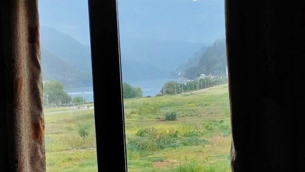 Udsigt gennem et vindue med gardiner, der viser et bjerglandskab, et vandområde og en bygning i det fjerne.