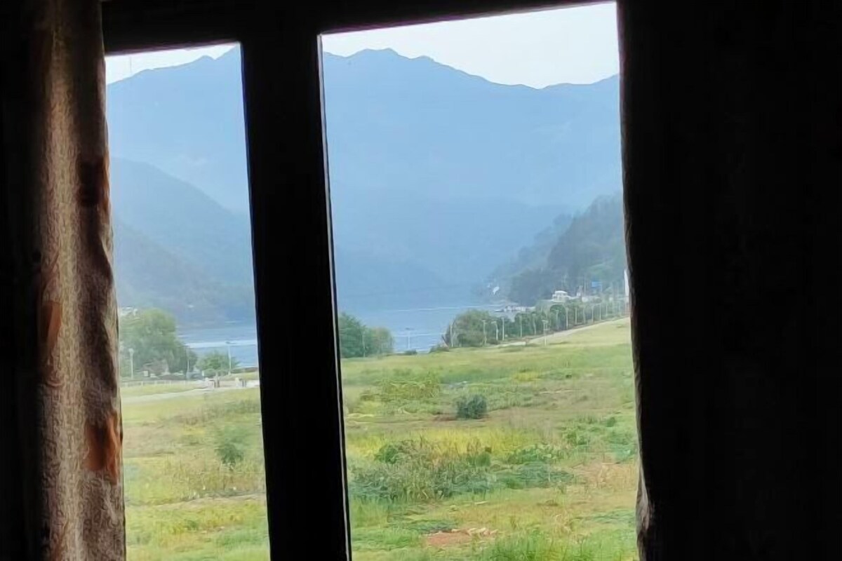 A view through a window with curtains, featuring a mountainous landscape, a body of water, and a distant building.