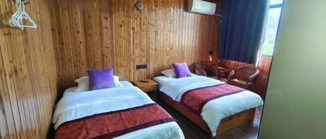 A room with two beds, wooden walls, a ceiling fan, a window with curtains, and a chair.