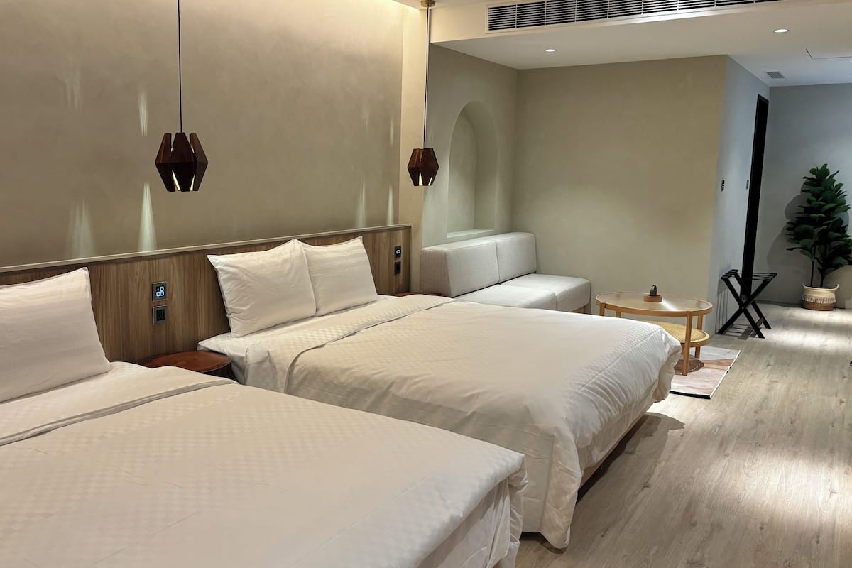 Ein Hotelzimmer mit zwei Betten, einem kleinen runden Tisch, einer Couch, einer Topfpflanze und einer wandmontierten Bedienkonsole.