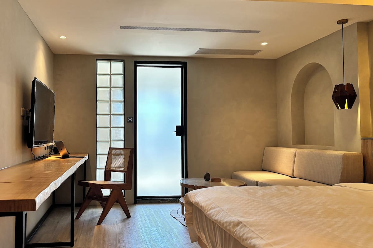 Ein Hotelzimmer mit einem Bett, einem Schreibtisch mit Laptop, einem Sessel, einem Fernseher und einem großen Fenster mit Milchglas.