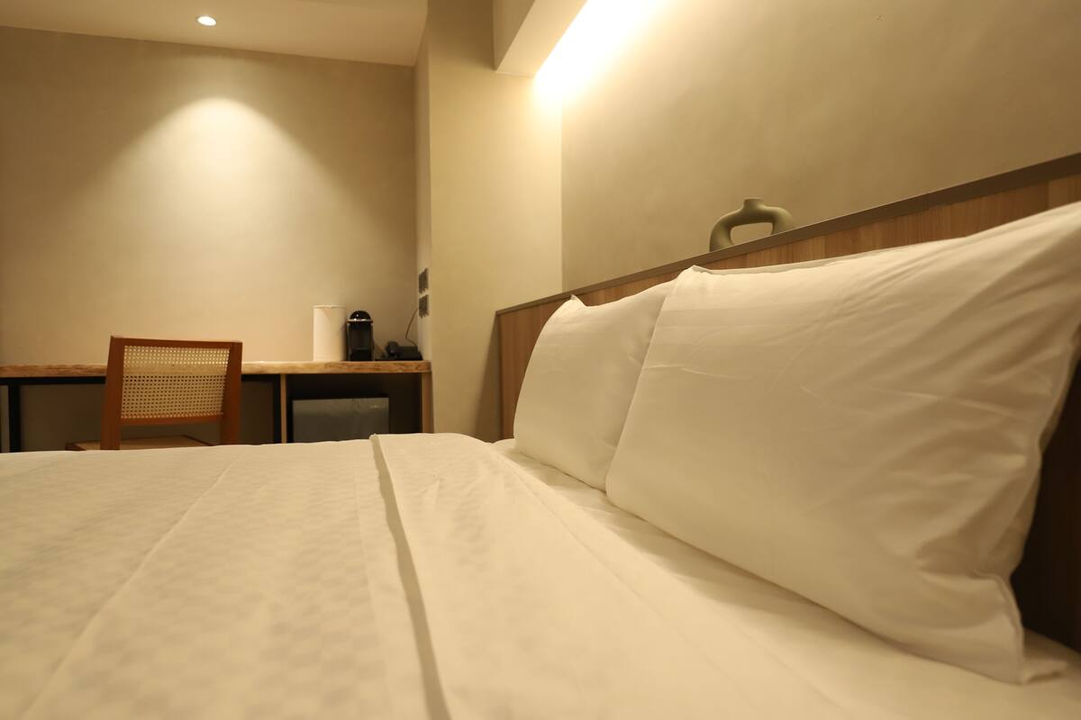 Ein Hotelzimmer mit einem ordentlich bezogenen Bett, einem Holzsessel, einem Schreibtisch mit Lampe und einem an der Wand befestigten Regal.