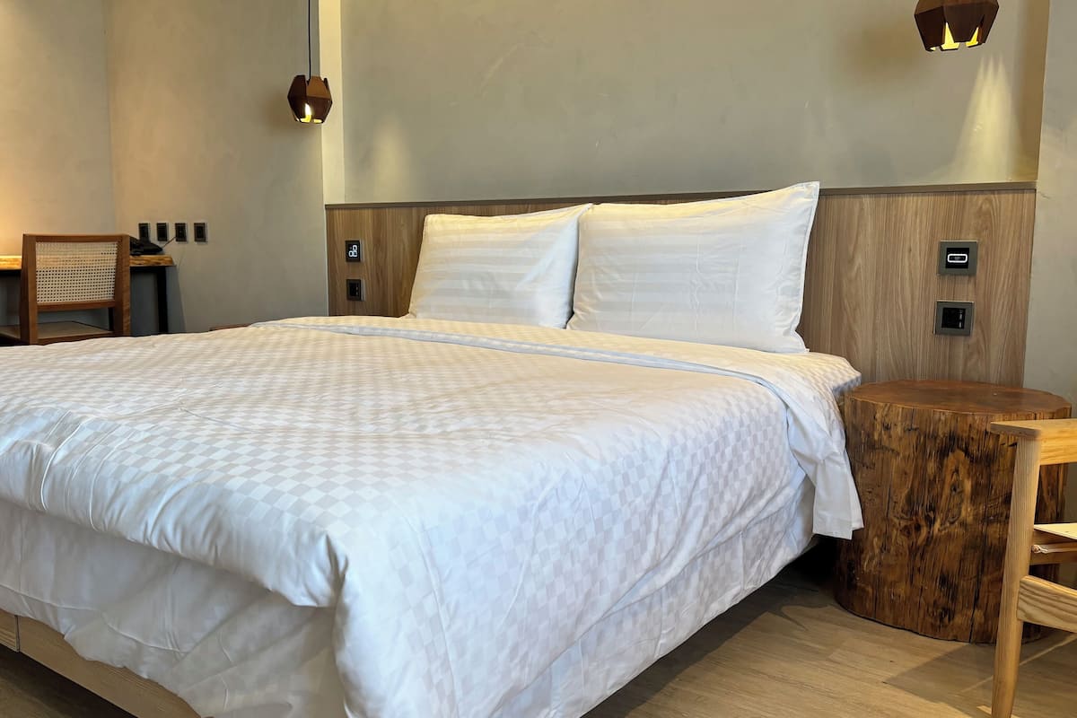 Ein ordentlich bezogenes Bett mit einer karierten Tagesdecke, einem Nachttisch aus Holz und einer an der Wand befestigten Lampe.