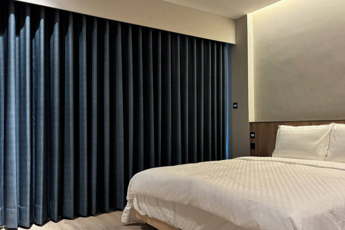 Ein modernes Hotelzimmer mit einem großen Bett, dunklen Vorhängen und einer neutralen Farbgestaltung.