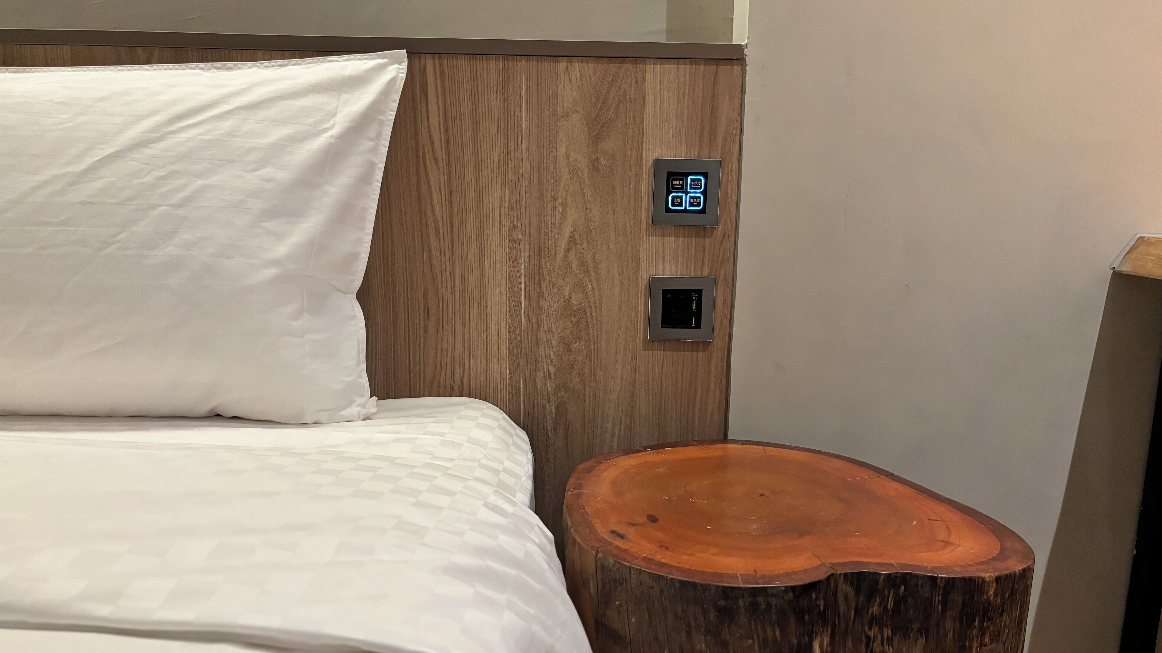 A neatly made bed with white linens, a wooden headboard, and two wall-mounted control panels.