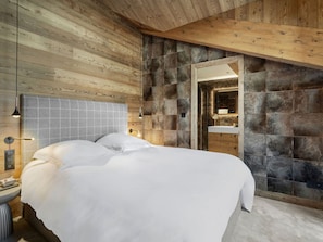 Quarto moderno com teto de madeira, uma cama grande com roupa de cama branca, parede de pedra e banheiro com espelho e pia.
