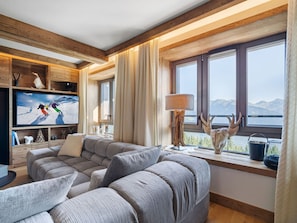 Uma sala de estar acolhedora com uma grande televisão de tela plana, um sofá modular e uma janela com vista panorâmica para as montanhas.