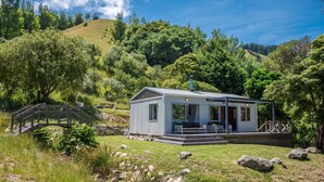 Une petite cabane moderne, dotée d’un porche et d’un escalier menant à l’entrée, entourée d’une végétation luxuriante et de collines.
